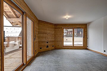 An empty room with wooden and white walls, a gray tiled floor, and two sets of sliding glass doors opening to snowy balconies and distant buildings.