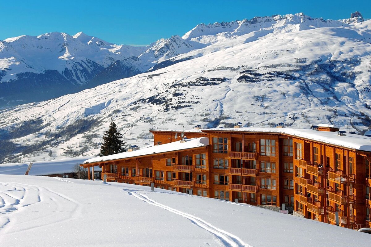 A large wooden ski lodge sits amidst a vast, snow-covered mountain landscape under a clear blue sky, with ski tracks in the foreground.
