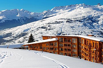 A large wooden ski lodge sits amidst a vast, snow-covered mountain landscape under a clear blue sky, with ski tracks in the foreground.