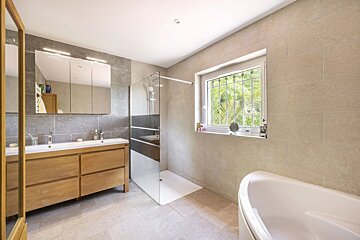 A modern bathroom with a wooden double vanity, walk-in glass shower, bathtub, and a window with security bars.
