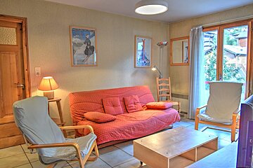 A cozy living room features a red sofa bed, two upholstered chairs, a coffee table, and ski posters. A large door opens to an outdoor view.