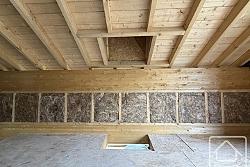 Interior of a wooden attic structure under construction, showing insulated walls, timber framing, ceiling rafters, and an attic hatch.