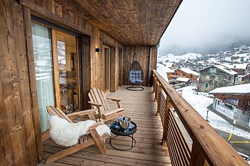 Cozy wooden chalet balcony with lounge chairs, overlooking a snowy alpine village.