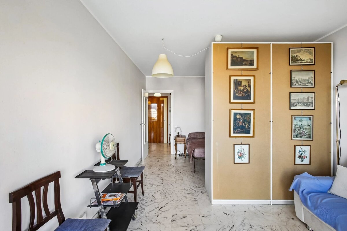 Bright, marble-floored room featuring a paneled room divider hung with pictures, a blue sofa, and a hallway leading to a bedroom.