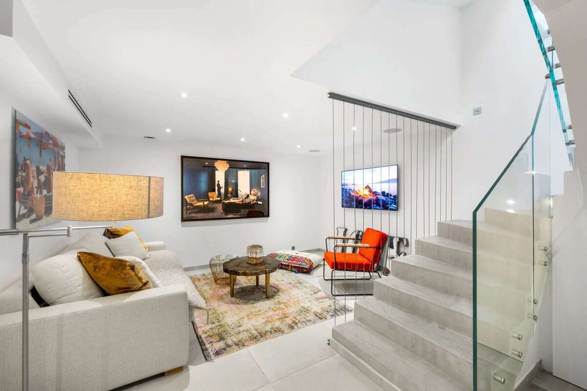 A stylish, modern living room with a white sofa, orange accent chair, colorful rug, large art, TV, and a chic glass-railed staircase.