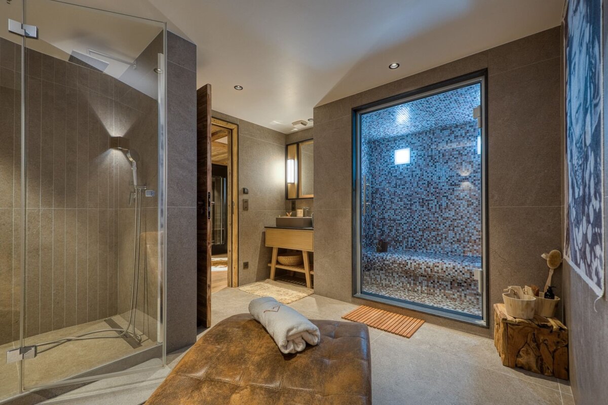 A modern spa bathroom featuring a glass shower, tiled steam room, vanity, and a leather massage bench with a towel.