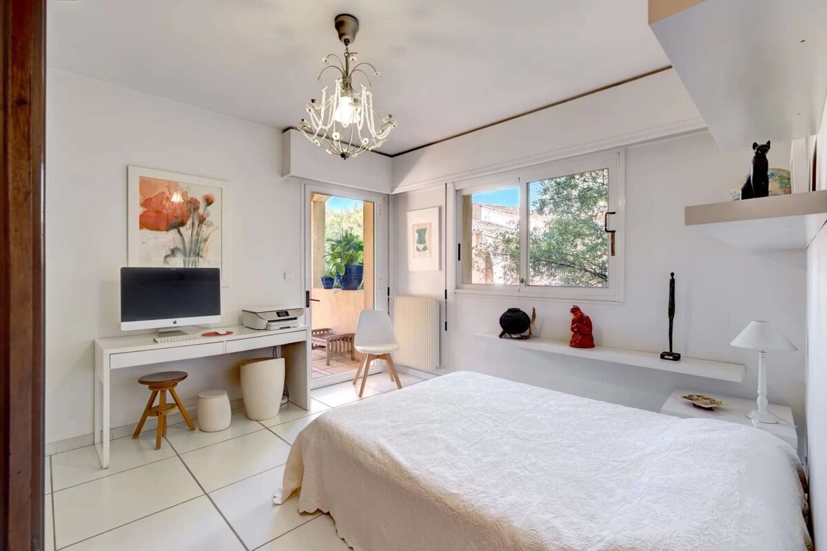 Bright, modern white room with a bed, desk, computer setup, and a door to a balcony. A chandelier and clean decor complete the space.