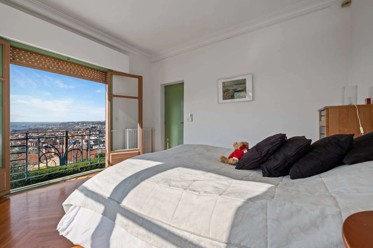 Bright bedroom with an open balcony revealing a stunning panoramic city and sea view. A bed with a teddy bear sits indoors.