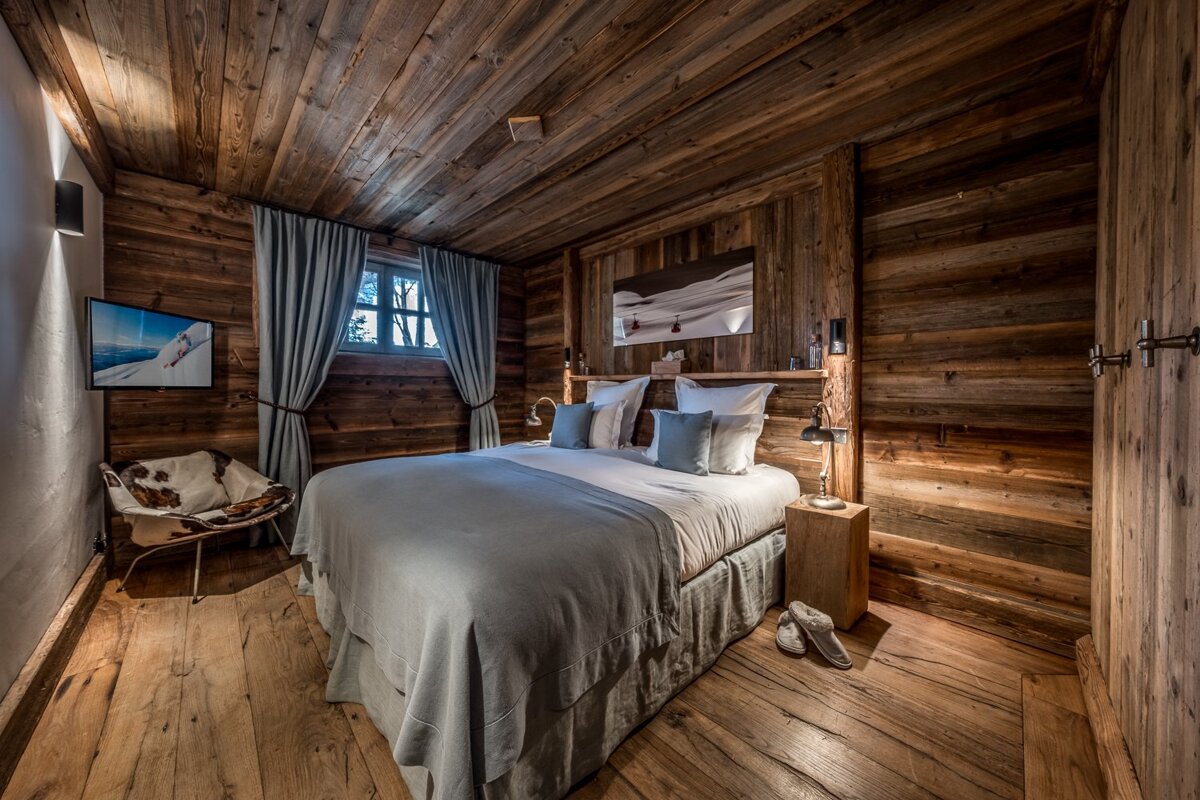 Cozy cabin bedroom with rustic wood-paneled walls and ceiling, featuring a bed with light blue linens, a window, and warm lighting.