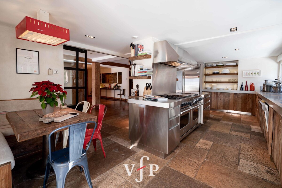 Rustic-modern kitchen with stone tile flooring, a stainless steel island stove, and a wooden dining table with colorful metal chairs.