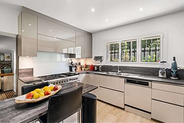 A modern kitchen features sleek beige cabinets, dark countertops, a stainless steel range, and a fruit bowl on a dark table. Three windows offer a view of greenery.