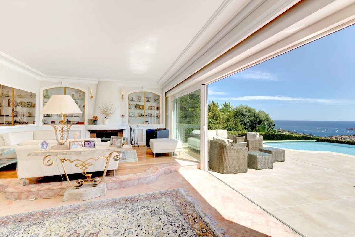 Bright living room with open glass doors overlooking a sunny patio, infinity pool, and a beautiful ocean vista.