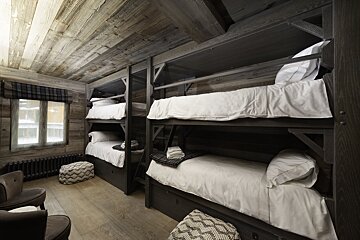 A cozy, rustic room features multiple dark wooden bunk beds with crisp white bedding. Distressed wooden walls, ceiling, and floor complete the cabin-like interior.