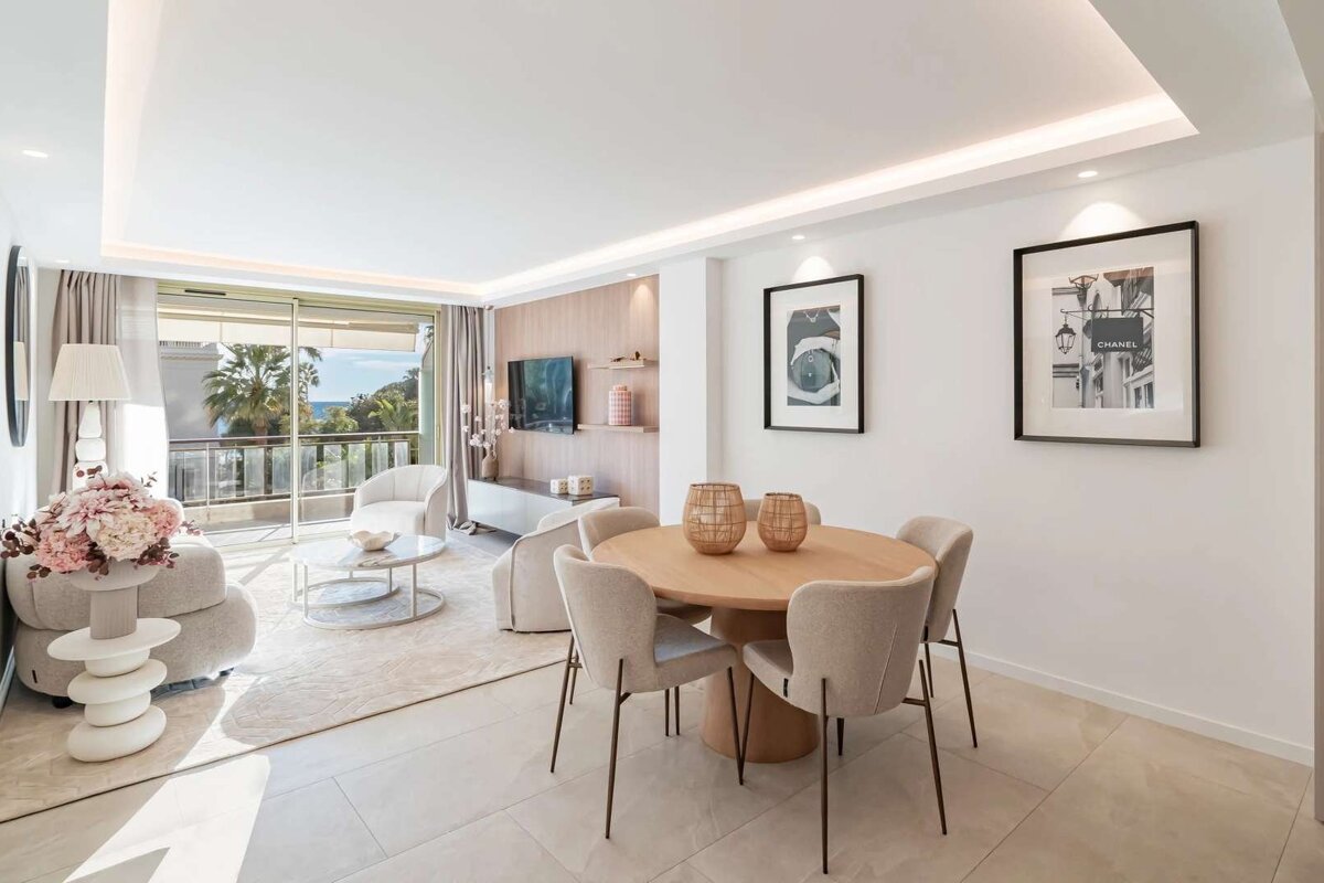 Elegant, neutral-toned living/dining room featuring modern decor, stylish furniture, and a balcony with a beautiful sea and palm tree view.