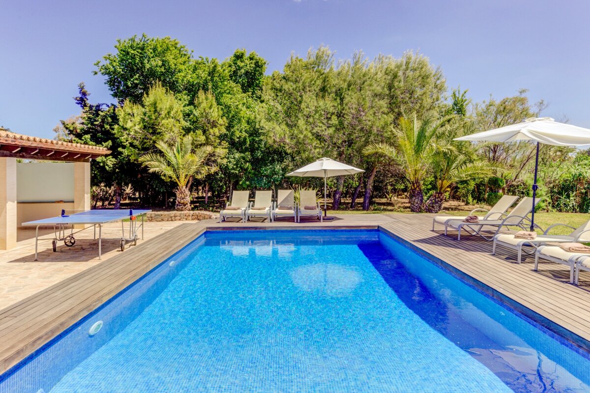 Bright outdoor pool area with a blue tiled pool, wooden deck, sun loungers, and lush green trees. A ping pong table sits nearby.