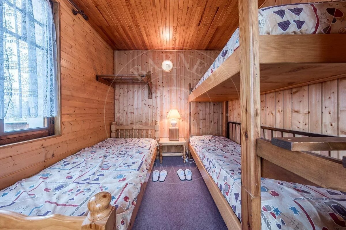 A rustic wooden bedroom with a single bed, bunk beds, patterned bedding, a nightstand with a lamp, and a window with lace curtains.