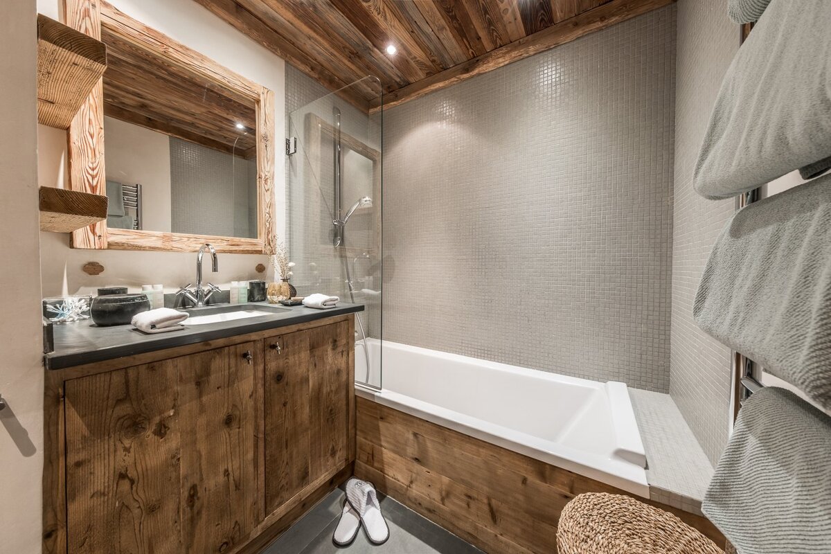 Cozy chalet-style bathroom featuring a wooden vanity, matching wood-paneled bathtub, grey tiled walls, and a warm wooden ceiling.