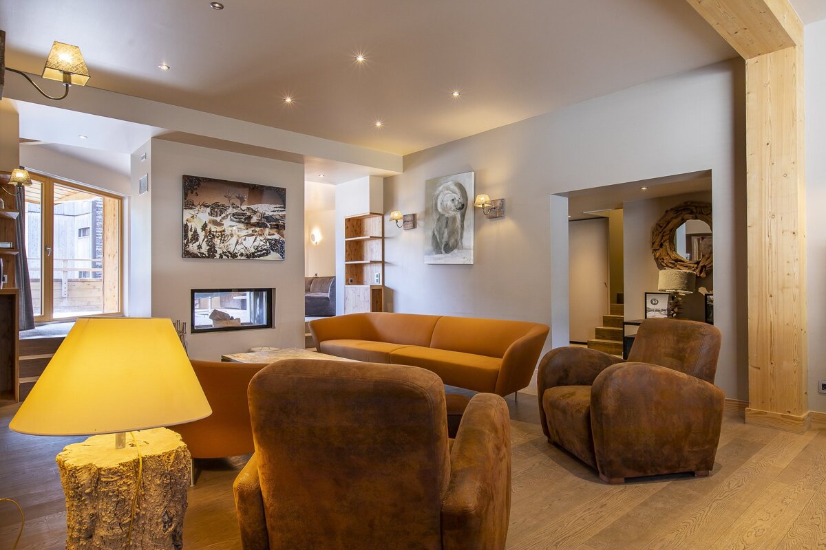 A cozy, modern chalet living room with an orange sofa, brown armchairs, fireplace, wooden decor, and warm lighting.
