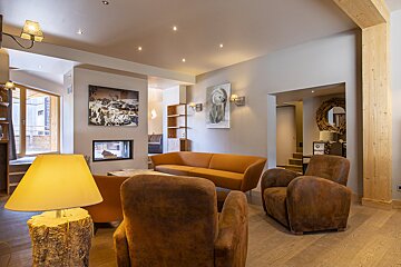 A cozy, modern chalet living room with an orange sofa, brown armchairs, fireplace, wooden decor, and warm lighting.