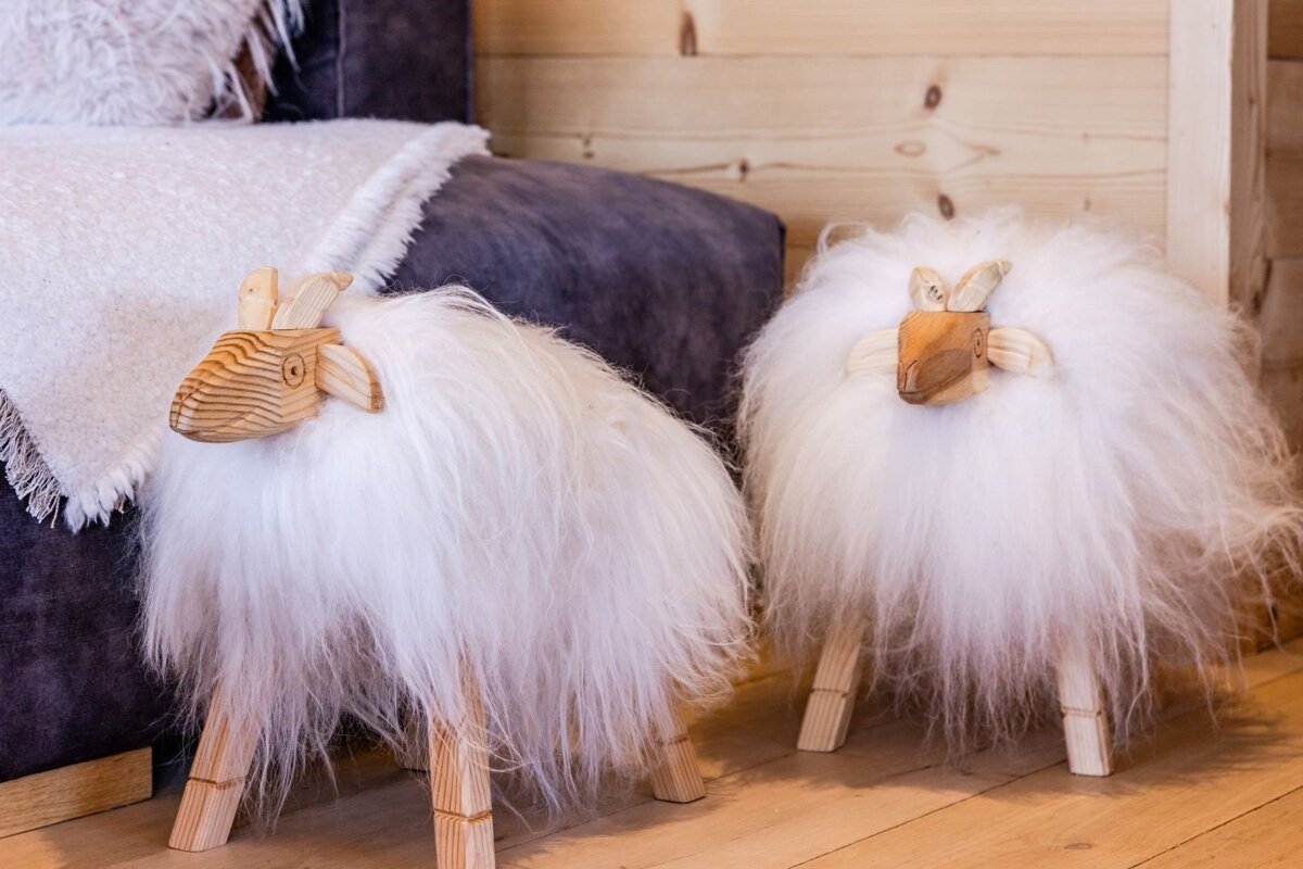 Two decorative wooden sheep with fluffy white wool standing on a wooden floor next to a couch.
