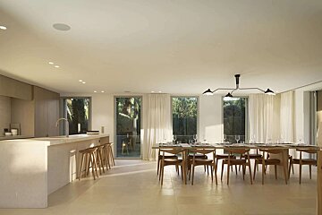 A bright, minimalist kitchen and dining space featuring a long island, a set dining table, and large windows with outdoor views.