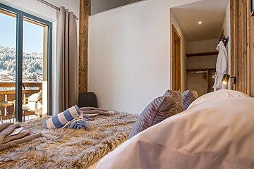 Cozy chalet bedroom with a fur-covered bed, scenic balcony views of snowy mountains, and an open closet.