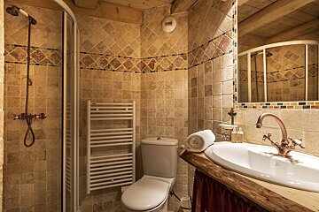 A rustic bathroom featuring earth-toned tiled walls, a corner shower, toilet, sink with a copper faucet, and a towel warmer.