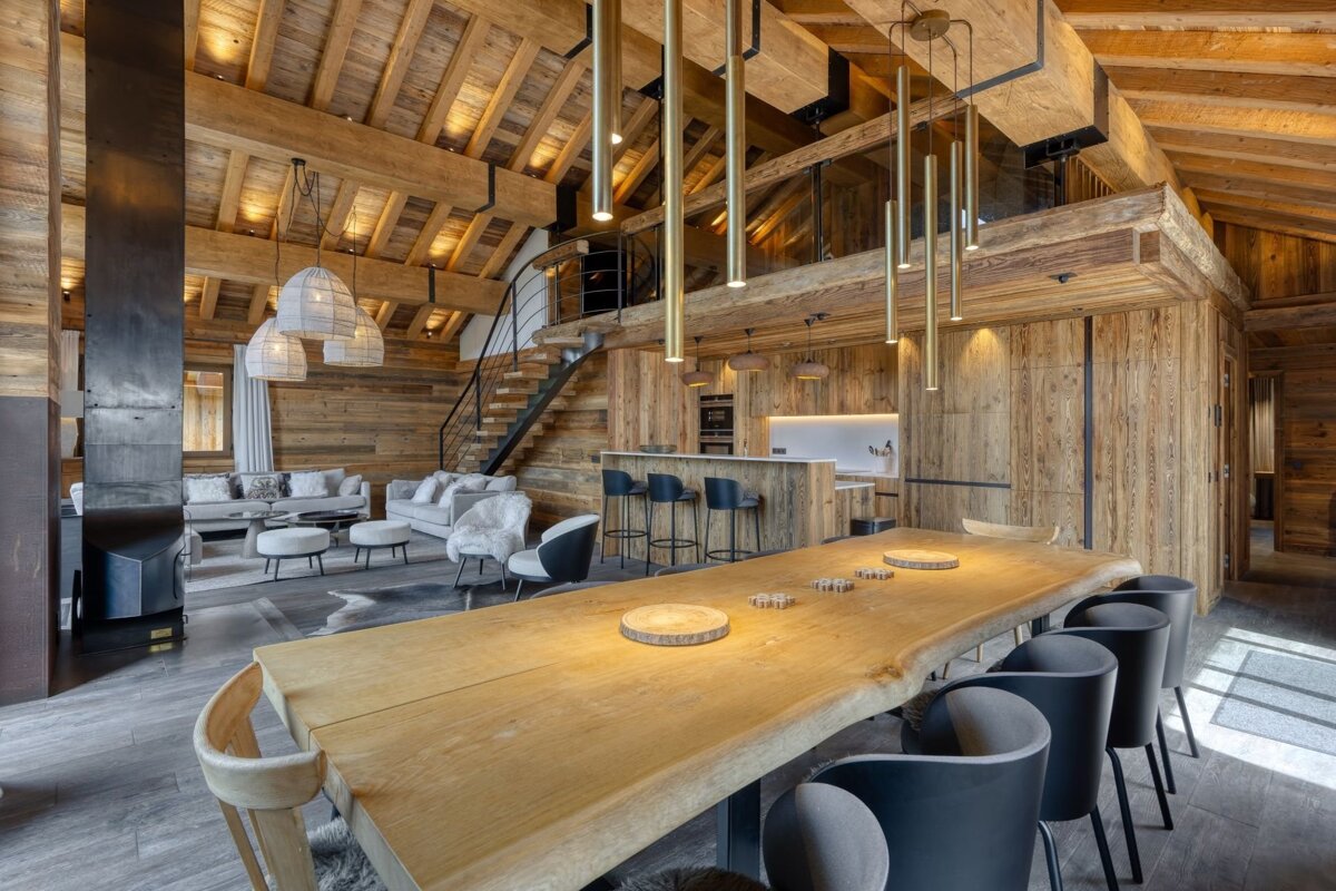 Luxurious, spacious open-plan chalet with abundant natural wood, modern furnishings, a long dining table, living area, kitchen, and mezzanine.