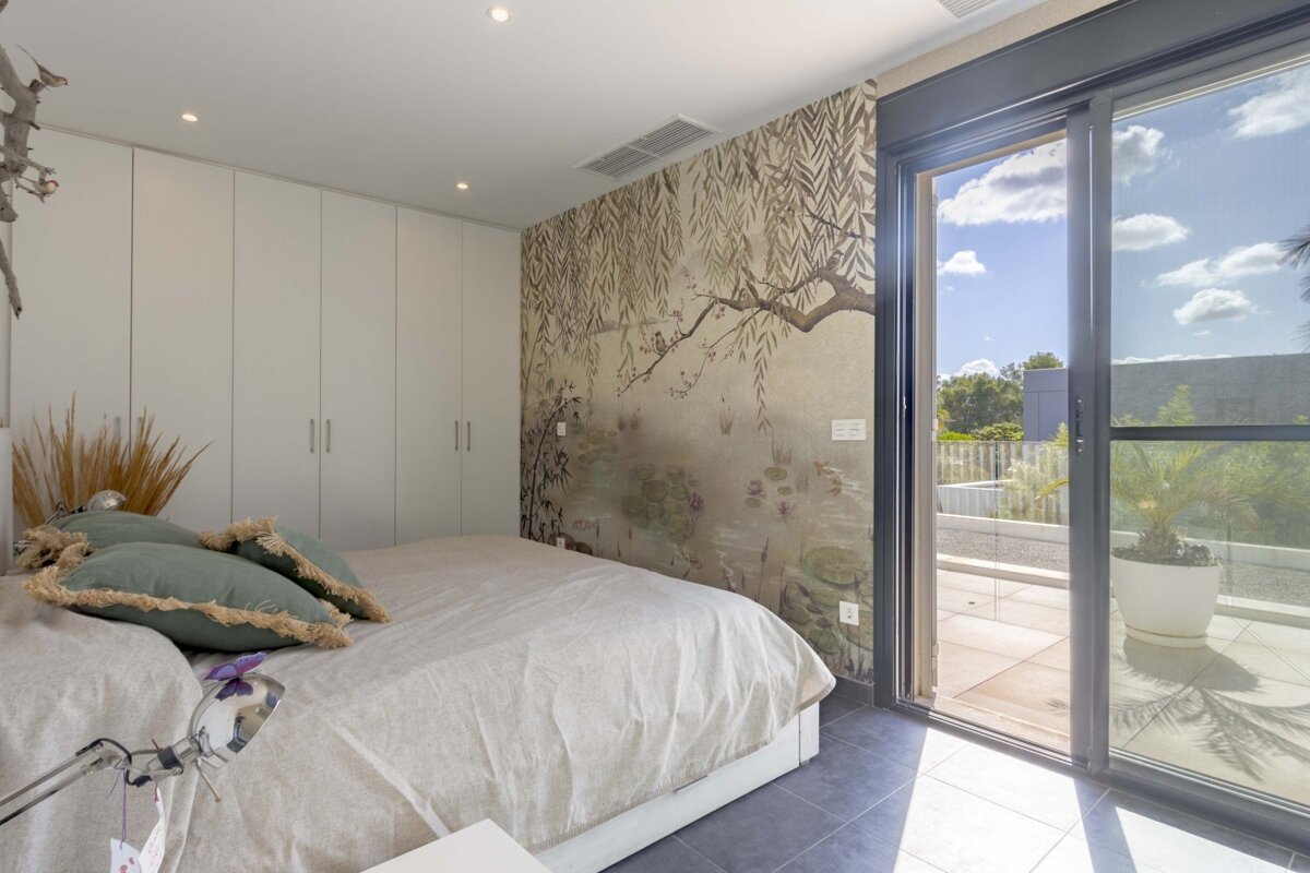 A modern bedroom features a bed with green pillows, white built-in wardrobes, and a nature-themed mural. A glass door opens to a sunny terrace.