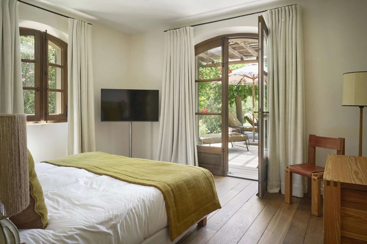 A serene bedroom featuring a white bed with a green throw, wooden floors, and a wall-mounted TV. French doors lead to a sunny outdoor patio with lounge chairs.