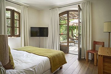 A cozy bedroom with a white bed, green blanket, TV, and French doors opening to a bright outdoor patio with lounge chairs and green foliage.