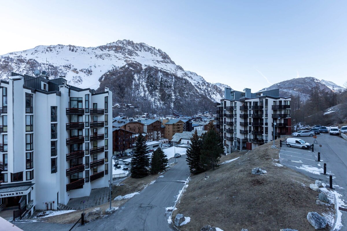 A mountainous ski resort town with multi-story buildings, snow-capped peaks, and scattered pine trees under a clear sky.