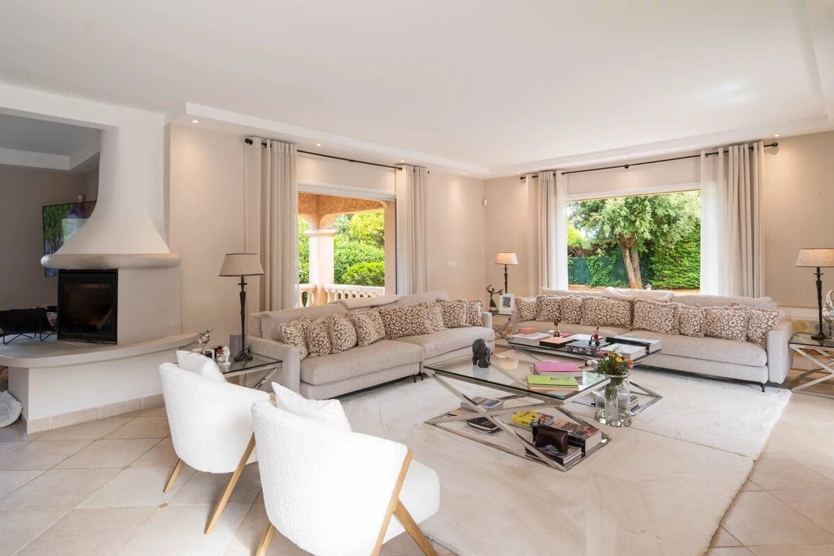 A bright, elegant living room featuring neutral-toned sofas, glass tables, white chairs, a fireplace, and large windows with garden views.