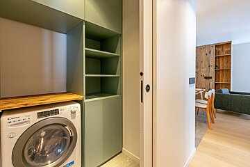 A laundry nook with a washing machine, wooden counter, and sage green built-in shelves. A white sliding door opens to a living area with wood accents.