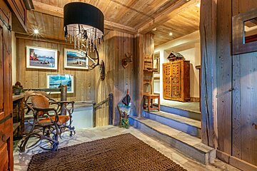 Cozy, rustic wooden chalet interior with stone floors, antler decor, a unique chandelier, an antler chair, and steps leading to a traditional cabinet.