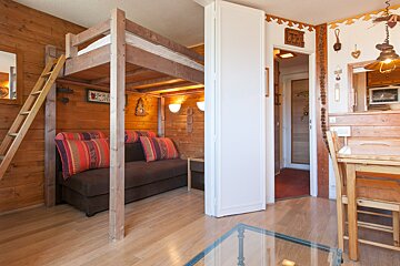 A cozy, rustic studio apartment with wood-paneled walls, a loft bed above a sofa, a dining area, and a white partition door.