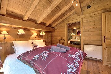 Cozy wooden chalet bedroom with an Edelweiss-patterned bed, teddy bear, and ensuite bath. Warm lighting creates a rustic, inviting atmosphere.