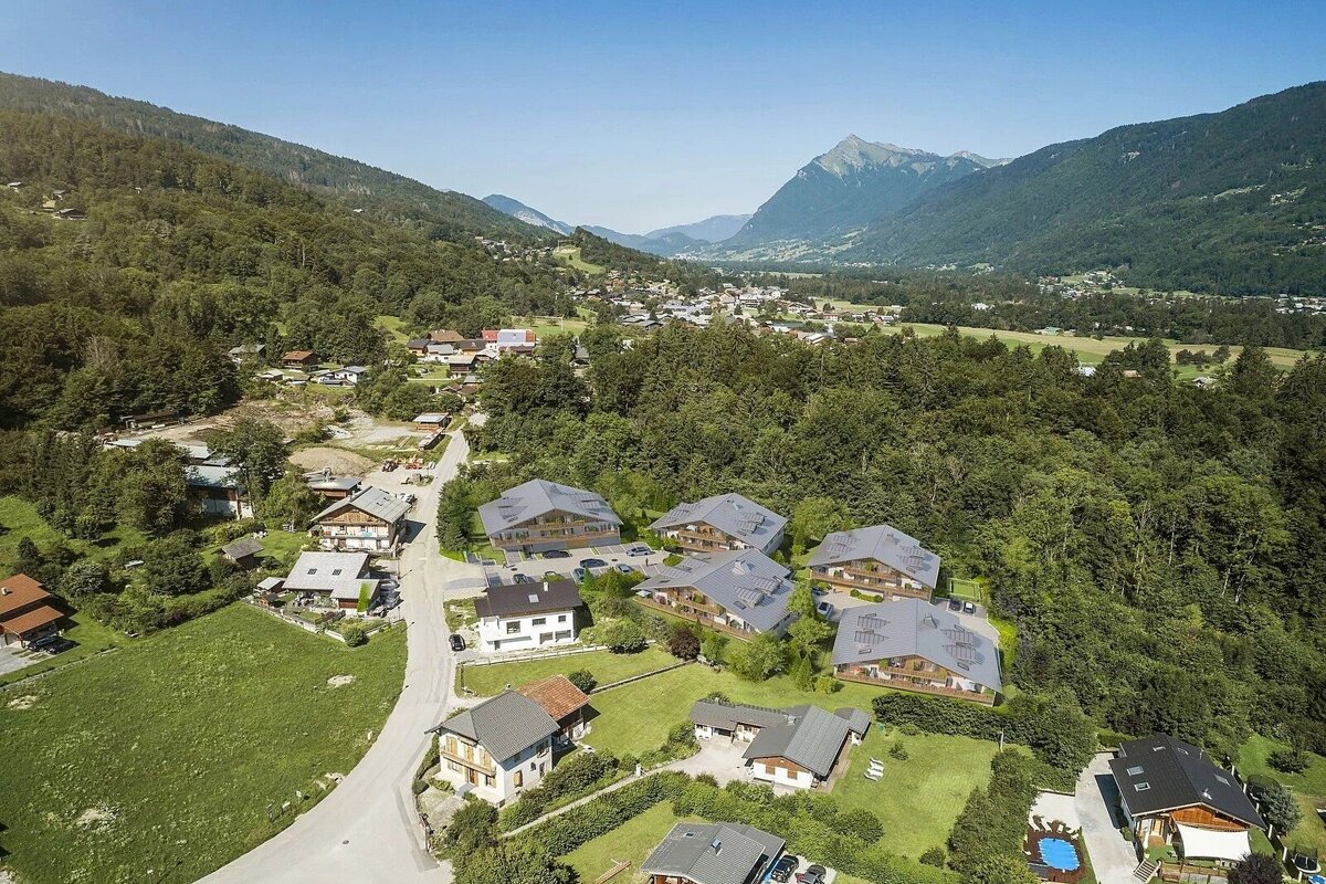 Aerial view of a mountain valley village with modern chalets nestled among green forests and distant peaks under a clear blue sky.