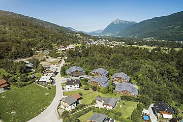 Aerial view of a mountain valley village with modern chalets nestled among green forests and distant peaks under a clear blue sky.