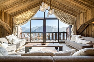 A cozy, wood-paneled chalet living room with large windows framing a stunning mountain vista. Sofas and a modern coffee table create a warm retreat.