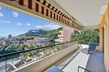 A sunny balcony with a lounge chair, covered by a striped awning, overlooking a coastal city with hills and buildings.