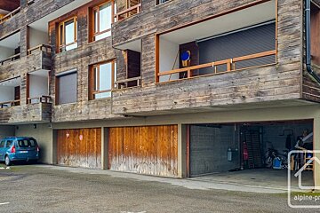 Weathered wooden apartment building with balconies, ground-floor garages (one open with bikes), and a blue car parked outside.