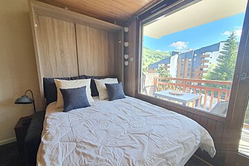 A room with a bed integrated into a wooden wall unit, a large window, and a balcony offering a view of mountains and buildings.