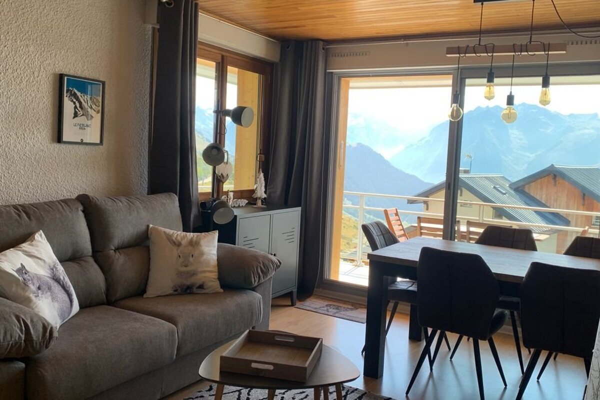Cozy living and dining area in a mountain cabin, featuring a brown sofa and a large glass door with a stunning mountain view.