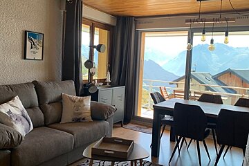 Cozy living and dining area in a mountain cabin, featuring a brown sofa and a large glass door with a stunning mountain view.