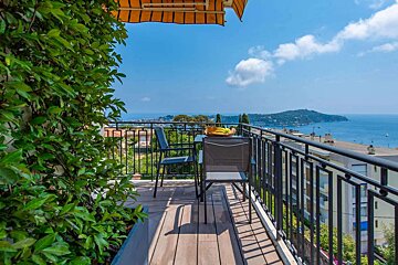 A sunny balcony with a table, chairs, and green foliage offers a stunning panoramic view of a blue ocean, distant coastline, and clear sky.