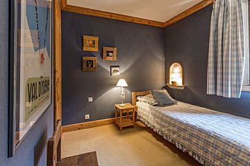 A cozy single bedroom with dark blue walls, wooden trim, a plaid bed, and a lit bedside table. A ski poster and framed animal art add a charming rustic touch.