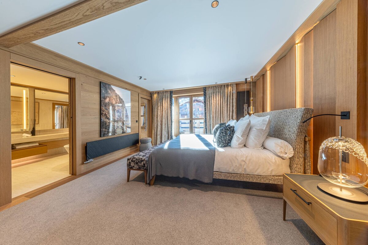 Elegant wood-paneled bedroom featuring a large bed, a modern glass bedside lamp, and a doorway leading into a brightly lit bathroom.