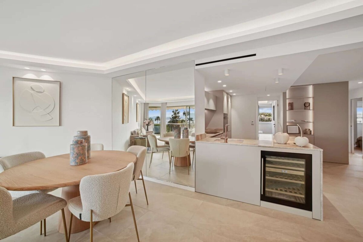 A chic, open-plan dining room and kitchen with light wood furniture, minimalist decor, and a mirrored wall reflecting a living area with an ocean view.
