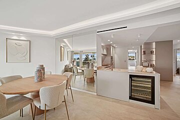 A modern, open-plan dining area with a wooden table, next to a sleek kitchen island with a wine fridge, all in light, neutral tones.